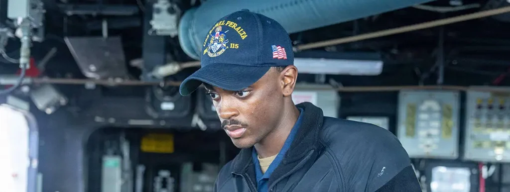 A focused U.S. Navy sailor wearing a USS Rafael Peralta (DDG 115) cap aboard a vessel, representing the personnel and operational readiness central to a potential US Blockade of Hormuz and the strategic complexities of the oil war.