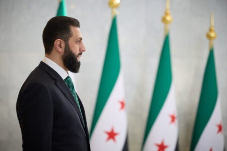 A profile view of Ahmad al-Sharaa in a dark suit standing before a row of official regional flags, representing the institutional context surrounding the implementation of the new Syrian investment law.