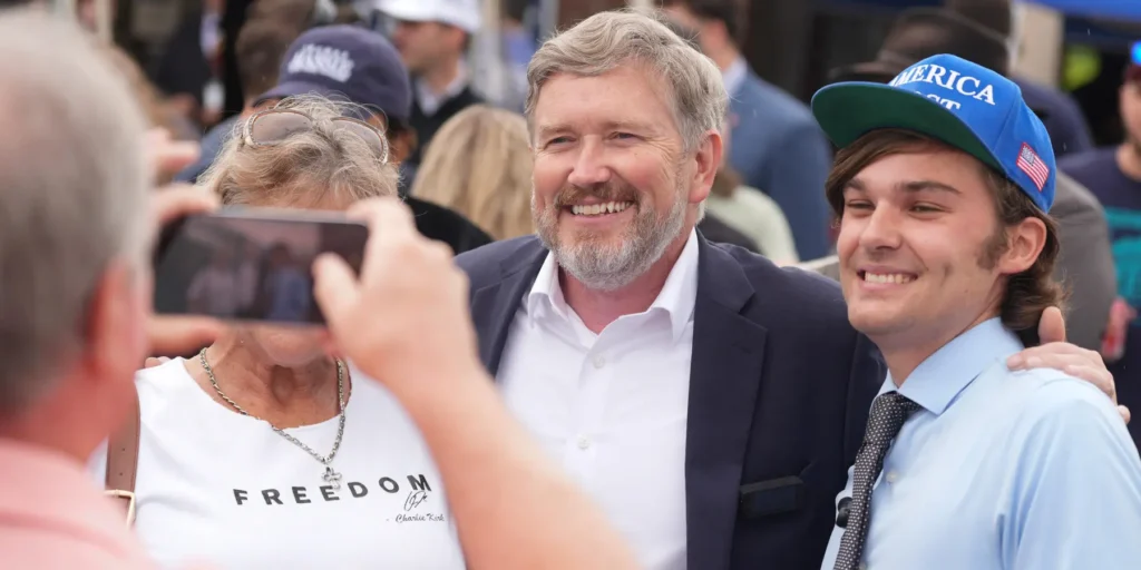 Representative Thomas Massie smiles while posing for a photograph with supporters wearing political attire, capturing the public engagement and grass-roots dynamics underlying his controversial 'America only' foreign policy stance.