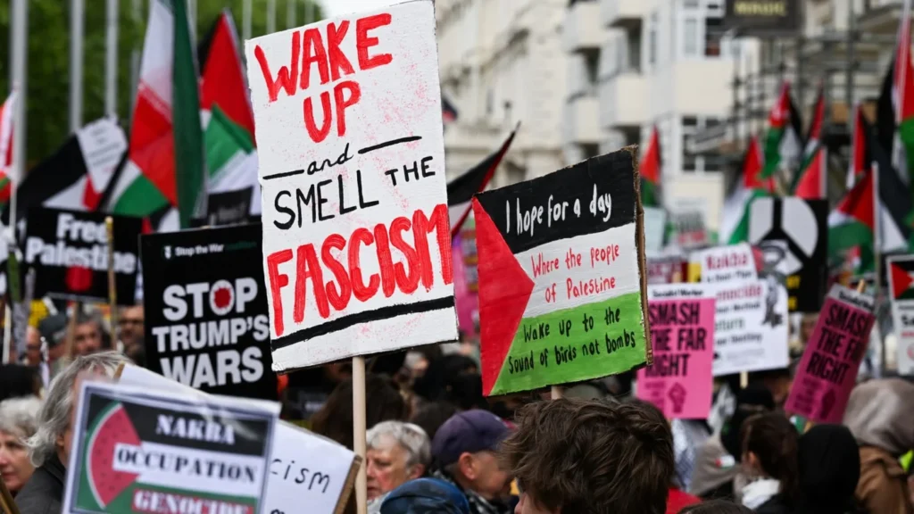 Demonstrators carry various political signs including one reading "Wake up and smell the fascism" during a crowded outdoor march, illustrating the societal tensions highlighted in the discussion From Nakba to fascist movements.