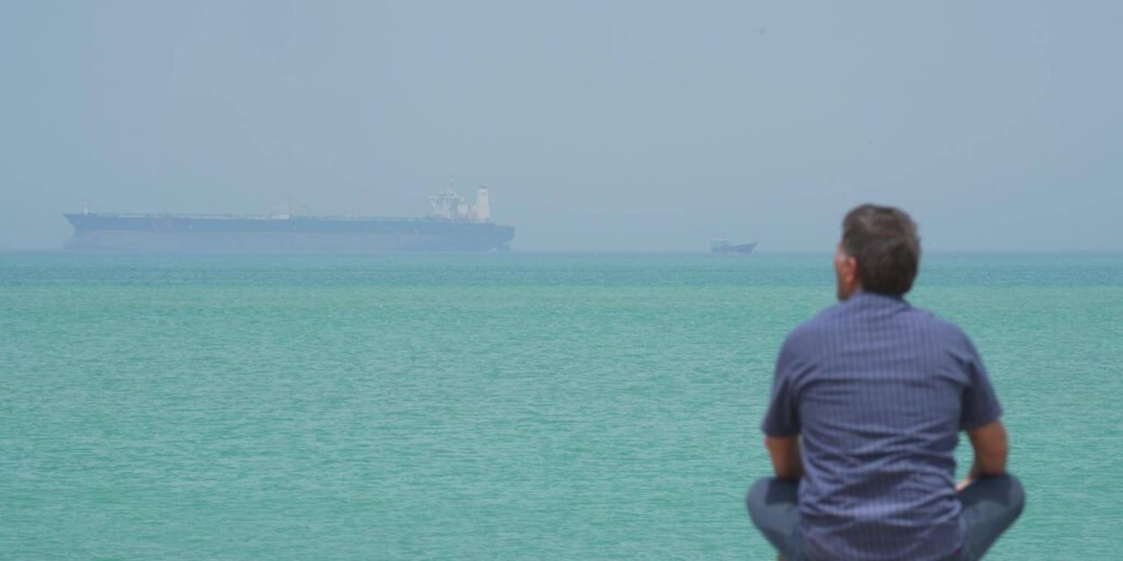 An observer watching a large oil tanker navigating the Strait of Hormuz, symbolizing the need for a naval coalition and regional maritime security.