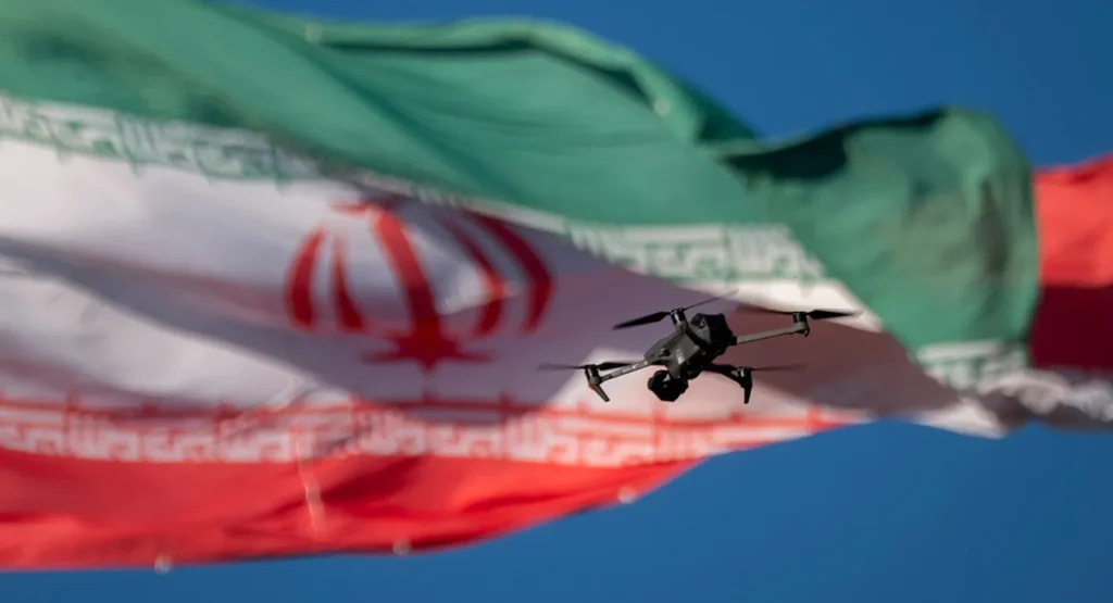 A close-up of a small tactical surveillance drone hovering in mid-air against the backdrop of a large, waving Iranian flag, symbolizing the technological shifts and high-tech strategies of Iran Asymmetric Conflict and the onset of AI-driven warfare.