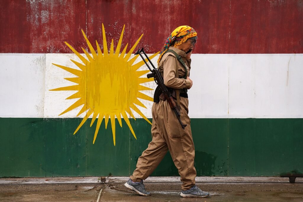 An armed Kurdish fighter walking past the Kurdistan flag, illustrating ethnic diversity and the potential for an Iranian Federation amid internal fractures.