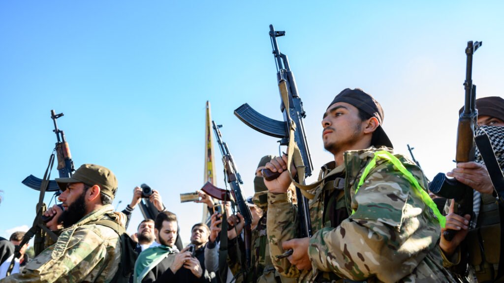 Armed personnel in camouflage attire hold up assault rifles during an outdoor gathering with onlookers under a clear sky, illustrating the persistent presence and ideological mobilization of the muhajirin within regional conflict zones.