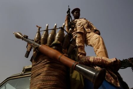 A low-angle shot of a soldier in desert camouflage uniform standing on a military vehicle next to a mounted rocket-propelled grenade (RPG) launcher and an array of warheads, illustrating the heavy militarization defining Sudan's civil war.