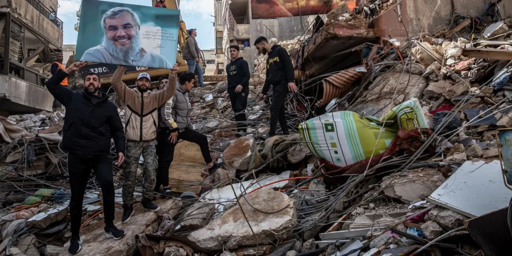 A group of men standing amidst the rubble of a destroyed building in a residential area, with two men holding aloft a large portrait of Hassan Nasrallah to signify Hezbollah strength during the 2026 peace talks.