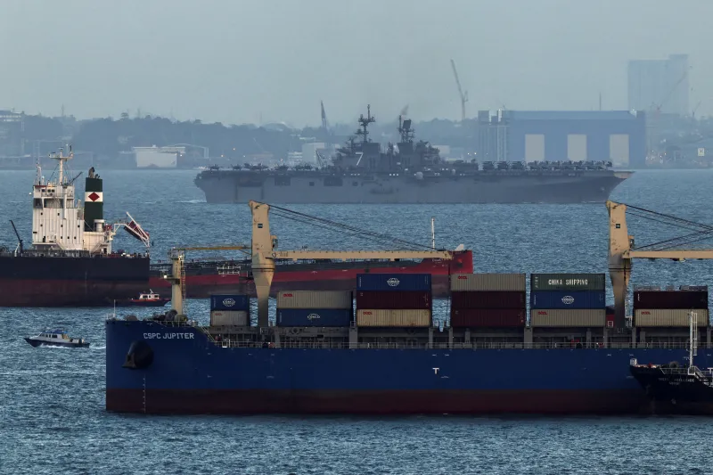 Hormuz Is a Warning for the Indo-Pacific A blue container ship named CSPC JUPITER operates in the foreground while a large military assault ship navigates the open waters in the background, showcasing the maritime security dynamics that explain why Hormuz Is a Warning.