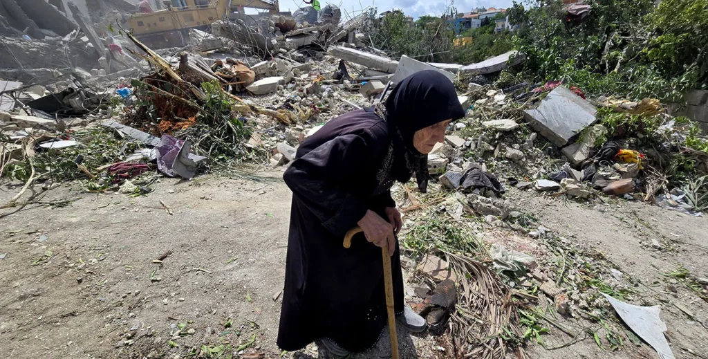 An elderly woman in a black dress uses a walking cane to navigate a wide path surrounded by massive concrete rubble, debris, and heavily damaged structures, illustrating the severe physical impact linked to Israel's Doctrine.