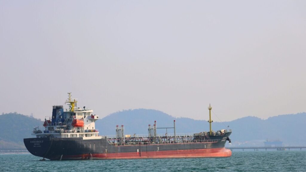 A commercial cargo vessel and oil tanker navigates open coastal water pathways near the Strait of Hormuz, highlighting the vulnerable transit infrastructure exposed to an economic polycrisis.