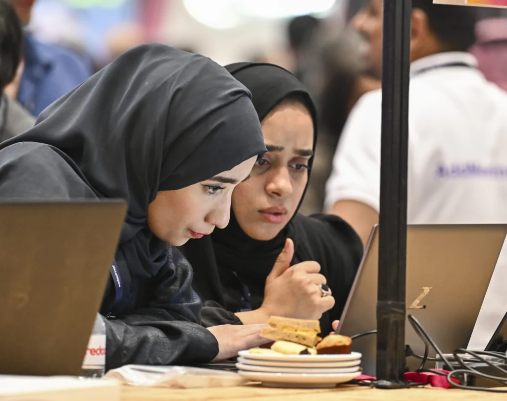 After the Iran war, the Gulf’s next economic phase awaits Two women wearing black hijabs look intently at a laptop screen during a technology conference, showcasing the digital innovation and workforce integration driving the Gulf’s next economic transformation.