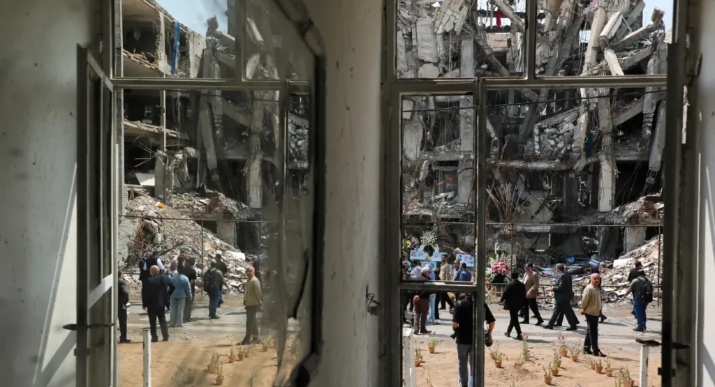 A view through a window frame of a heavily damaged building in Tehran following military strikes, with debris and structural ruins in the background, serving as a stark visual for the geopolitical consequences surrounding the Iran nuclear program and the aftermath of the Twelve-Day War.