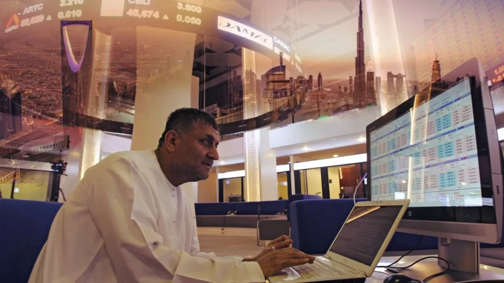 A financial analyst works at a dual-monitor trading desk displaying market data, with a digital overlay of the Dubai skyline and stock tickers, illustrating how War Pressures Gulf Economies behind closed doors.