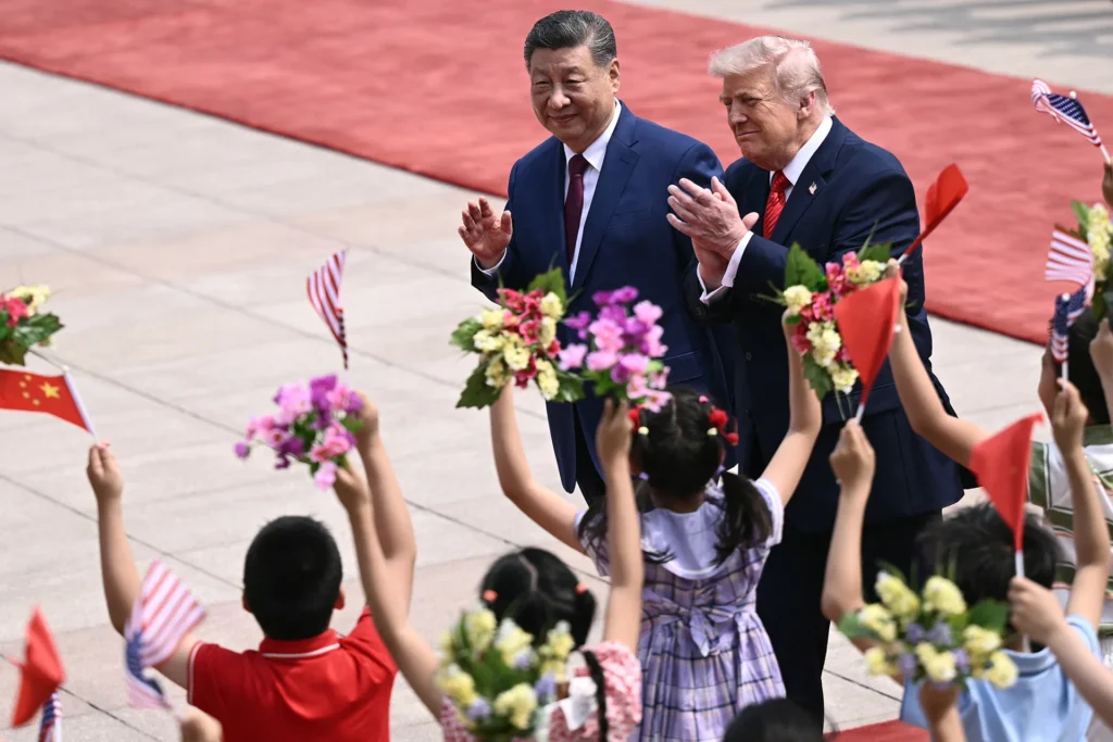 ما بعد تايوان: “سلام لائق” في قمة ترامب-شي Xi Jinping and Donald Trump wave and clap on a red carpet while children wave mini Chinese and U.S. flags, capturing the public diplomatic stage and underlying geopolitical balancing act of U.S.-China strategic stability.