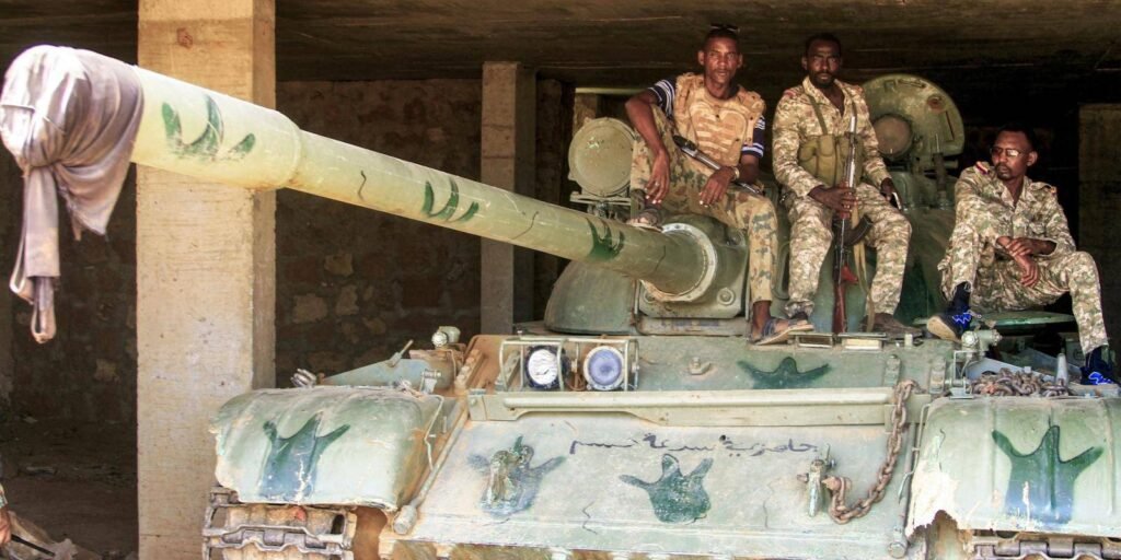 يمكن قطع تدفق الأسلحة والأموال التي تغذي الحرب في السودان. ما ينقص هو الإرادة Three military personnel in camouflage uniforms sit positioned atop a large, weathered combat tank parked inside a concrete structure, representing the local military control factors shaping the war in Sudan.