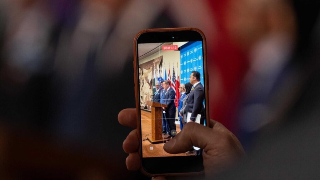 A hand holding a smartphone captures a video recording of Ambassador Michael Waltz of the USA speaking at a joint press briefing alongside Bahraini Ambassador Jamal Fares Alrowaiei at the UN Headquarters, focusing on regional cooperation and Gulf politics.