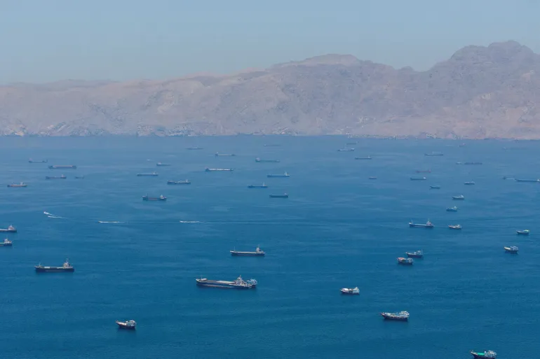 The strait may reopen, but global confidence may not return A wide high-angle view of numerous cargo ships and maritime vessels anchored or moving across the Strait of Hormuz with a barren mountainous coastline in the background, highlighting the supply chain vulnerabilities impacting global confidence.