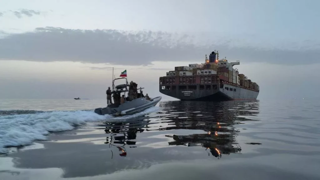 A fast-moving military interceptor boat flying the Iranian flag approaching a massive commercial container ship at sea, illustrating the high-stakes maritime tensions and escort protocols central to a potential U.S. Naval Blockade and the ongoing Iran standoff.