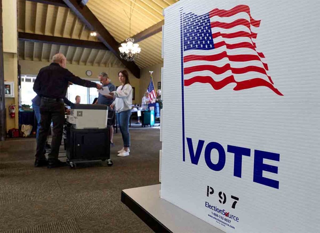 A close-up of a prominent white voting sign featuring the American flag and the word "VOTE," with individuals casting ballots in a blurred polling station backdrop, contextualizing the international influence and geopolitical weight linked to Arab Dependency on US.