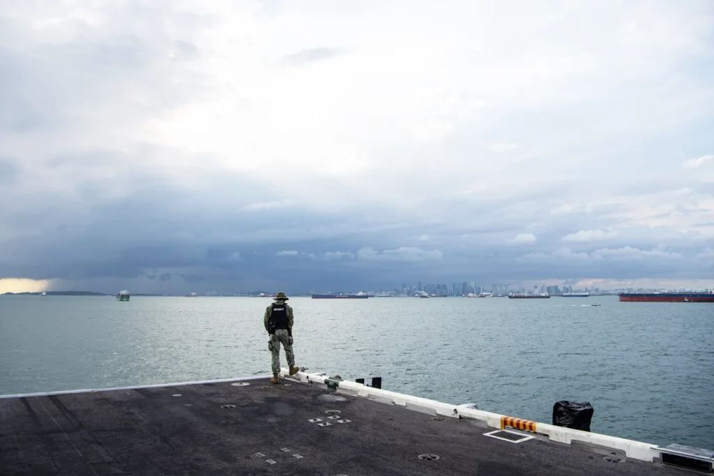A U.S. Navy sailor in full gear, Master-at-Arms Third Class Elisha Francois, stands watch on the deck of a Nimitz-class aircraft carrier as it transits the Strait of Malacca, symbolizing the high-stakes security measures and strategic Global Maritime Leverage required to navigate the world's most critical chokepoints.