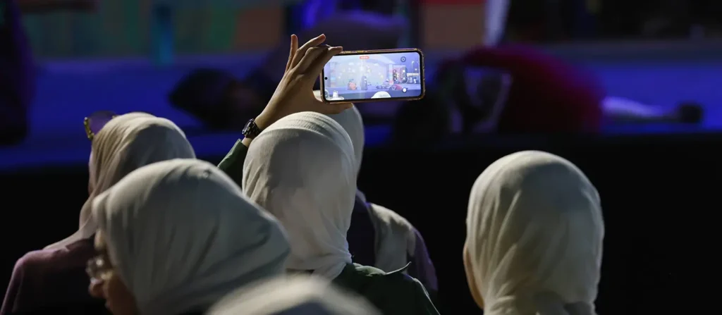 A woman in a crowd holding up a smartphone to record a public event, highlighting the mobile connectivity and user-level Syria Digital Infrastructure that remains a strategic priority for regional communication and stability.