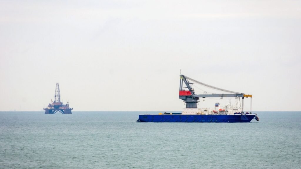 A blue industrial crane vessel and a distant offshore oil rig operating on the horizon, highlighting the energy infrastructure and maritime logistics essential for successful Caspian Sea Trade despite regional geopolitical tensions.