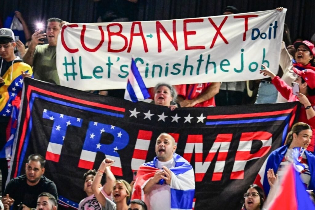Demonstrators hold a large banner reading "CUBA NEXT Do it #Let's Finish the Job" alongside a prominent Trump flag during a rally, highlighting public sentiments surrounding potential military intervention in Cuba.