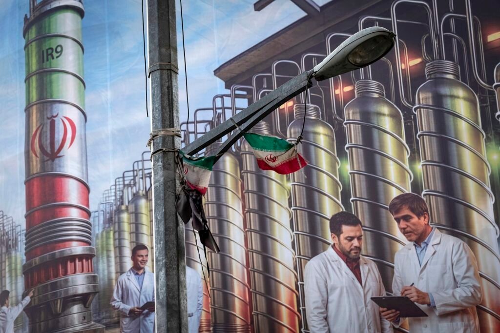 A street-level view of an Iranian mural featuring nuclear scientists and industrial IR-9 centrifuges, framed by a weathered national flag on a lamp post, symbolizing the regional tensions and geopolitical stakes inherent in modern Nuclear Weapons Strategy and the NPT framework.