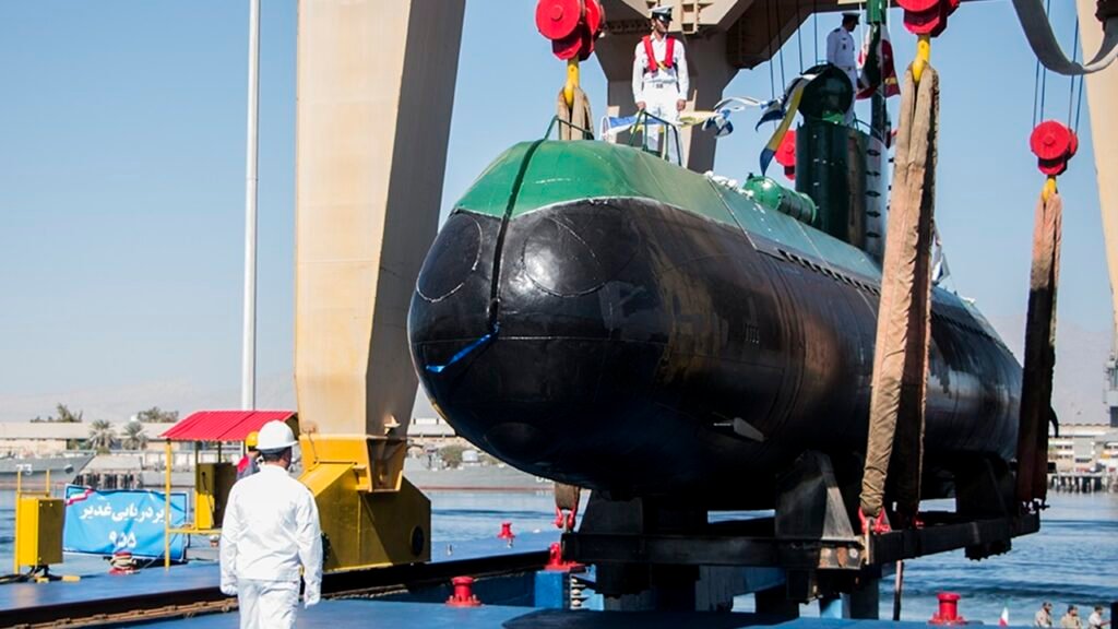 A small, black and green Ghadir-class diesel-electric submarine held in heavy lifting slings by a harbor gantry crane during a naval ceremony, showcasing the compact underwater assets that serve as a tactical focus for asymmetric warfare in shallow waters.