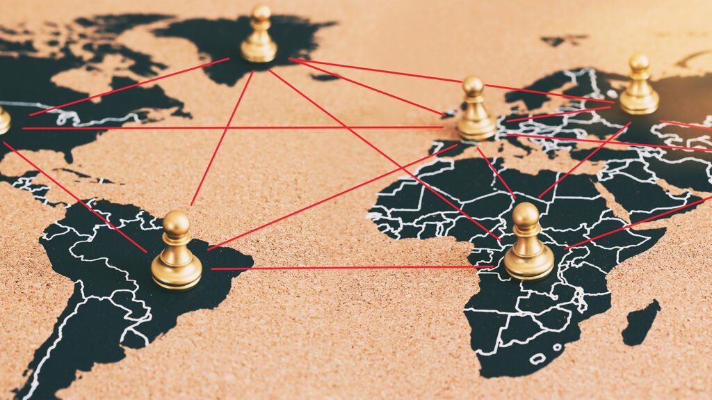 A cork world map with golden chess pawns placed on different continents connected by intersecting red lines, visually representing global networks and the geopolitical shifts foundational to a Decolonial Consensus.