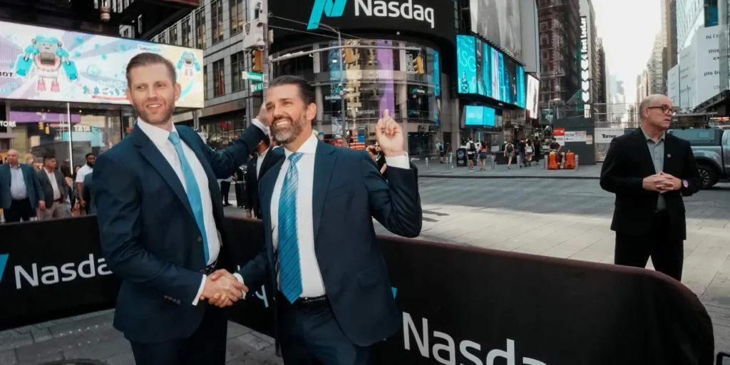 Donald Trump Jr. and Eric Trump in suits at Times Square with the Nasdaq sign in the background, symbolizing the intersection of high-finance, political influence, and the controversial discussions surrounding Iran War Grifting and war profiteering.