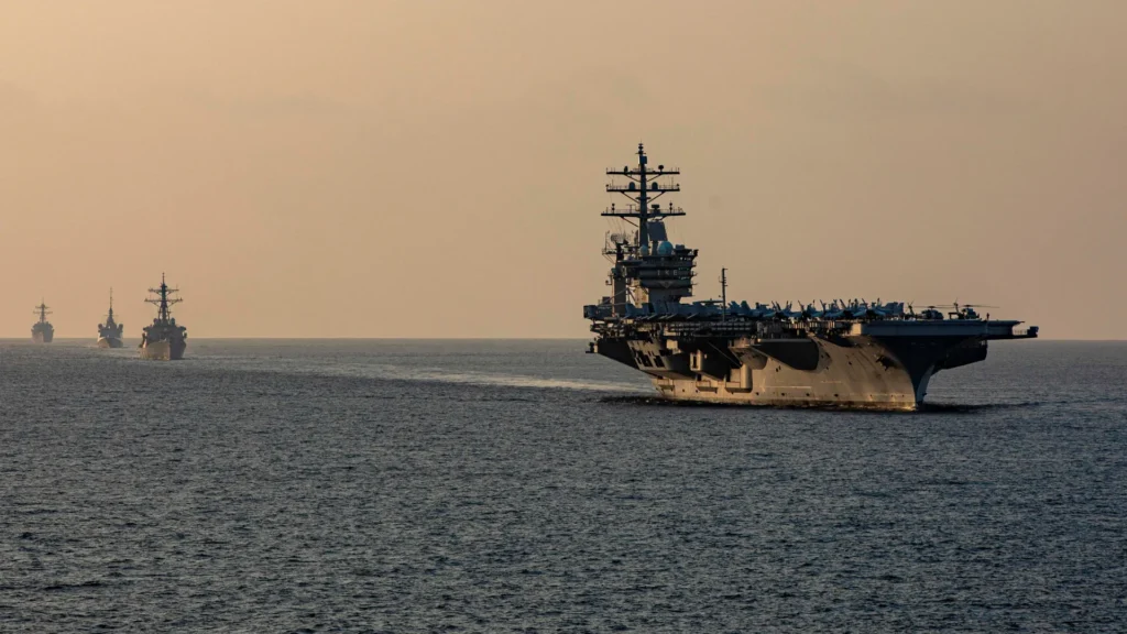 The USS Dwight D. Eisenhower aircraft carrier leads its strike group vessels through the open waters of the Strait of Hormuz, providing a visual focal point for evaluating the global economic and naval stakes of the Iran war.