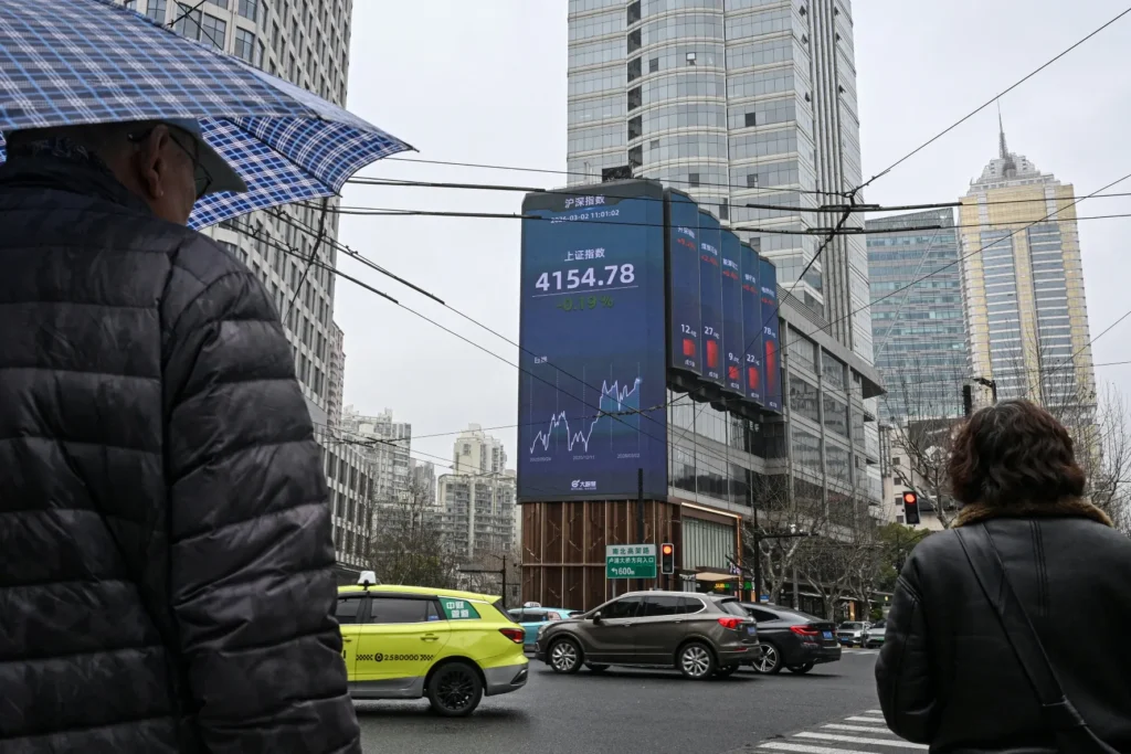 Pedestrians in a modern Chinese city passing a large digital billboard displaying stock market fluctuations and financial data, illustrating the economic vulnerabilities and market volatility tied to the China Iran Conflict and its impacts on global strategy.