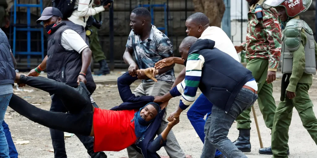 Several men desperately carry an injured demonstrator in a red shirt away from an active clash, while uniformed police officers advance in the background, capturing the volatile environment of regional Diesel protests.