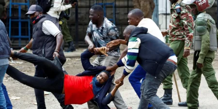 Several men desperately carry an injured demonstrator in a red shirt away from an active clash, while uniformed police officers advance in the background, capturing the volatile environment of regional Diesel protests.