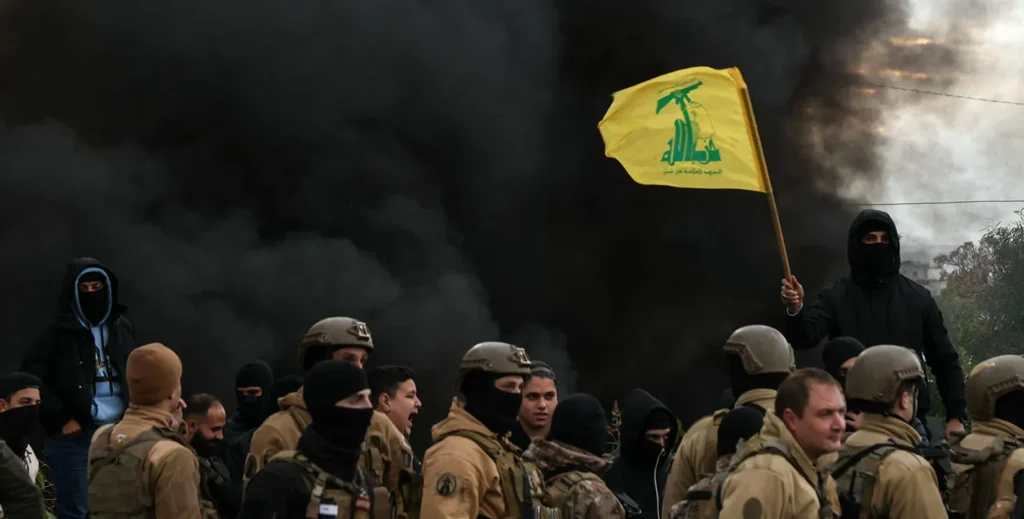 Pushing Beirut into an Armed Conflict With Hezbollah Is Insane A crowd of armed personnel in military gear and balaclavas gathers before a dense plume of black smoke, while an individual raises a yellow flag, underscoring the regional tensions and risks of an armed conflict with Hezbollah.