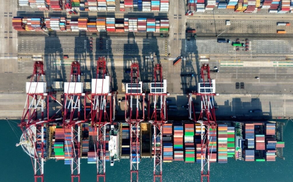 An aerial top-down view of a bustling container terminal with massive red cranes loading a cargo ship, representing the logistical infrastructure and supply chain resilience vital to Global energy security and revolutionary economic shifts.