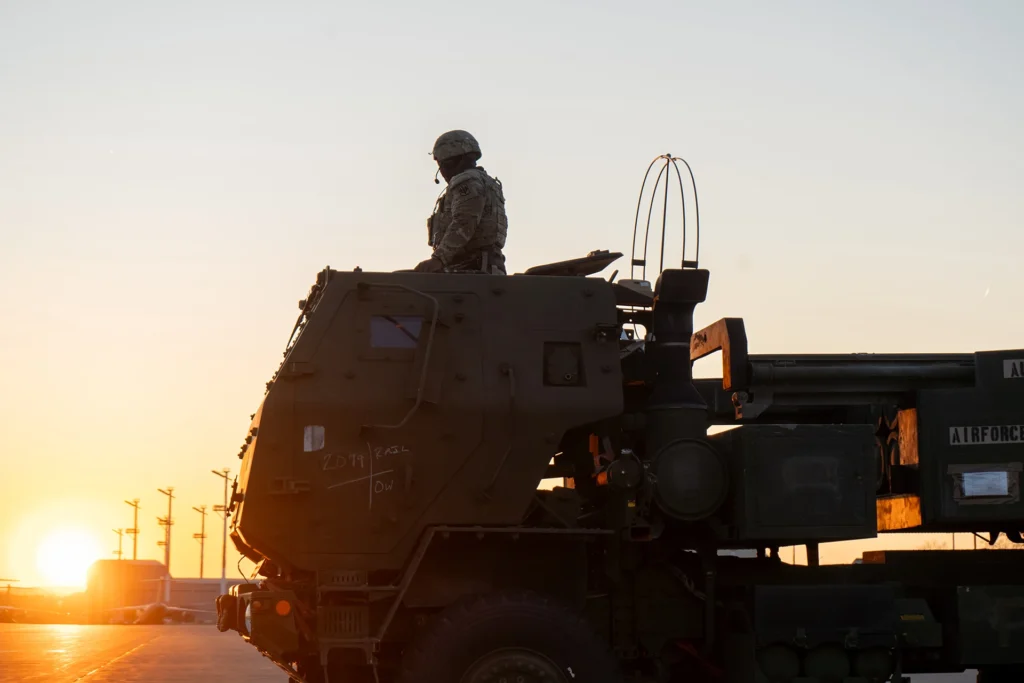 A silhouette of a soldier standing atop a military rocket launcher vehicle (HIMARS) during sunset at an airfield, symbolizing the defensive readiness and hardware deployment central to the NATO Deterrence Crisis and regional security challenges.