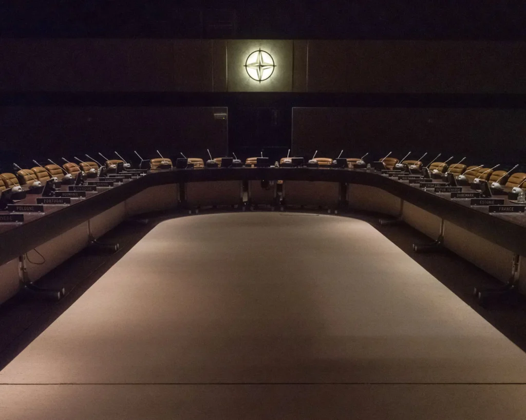 An empty, horseshoe-shaped conference table lined with microphones sits beneath a dimly lit NATO emblem in a dark boardroom, capturing the institutional vacuum and deliberation surrounding the NATO Middle East strategy.