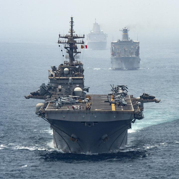 How to Accomplish U.S. Objectives in Iran A massive U.S. Navy amphibious assault ship carrying helicopters and military equipment leads a fleet across the open ocean, visualizing the naval presence and strategic military posture behind current U.S. Objectives in Iran.