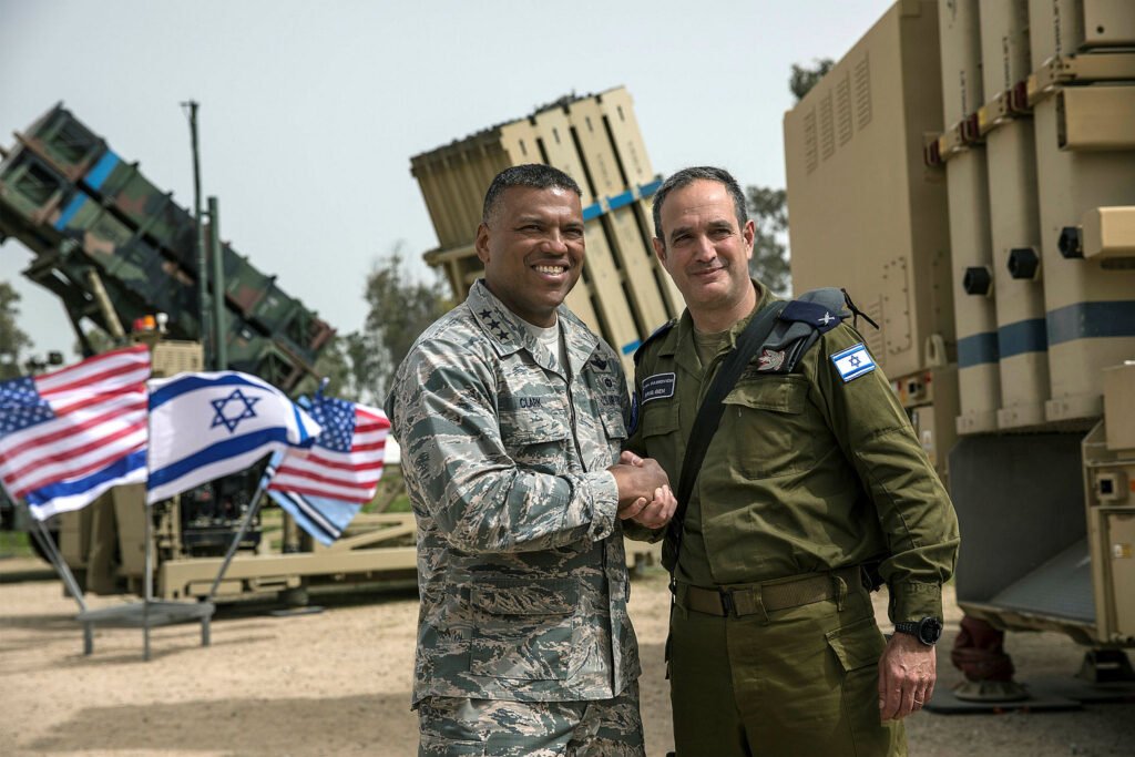 U.S. Air Force Lieutenant General Richard Clark shaking hands with Israeli Brigadier General Zvika Haimovich at a military base, with an Iron Dome launcher and a Patriot missile battery in the background, highlighting the defense partnership as the U.S. Marines move forward with Israeli interceptor technology.