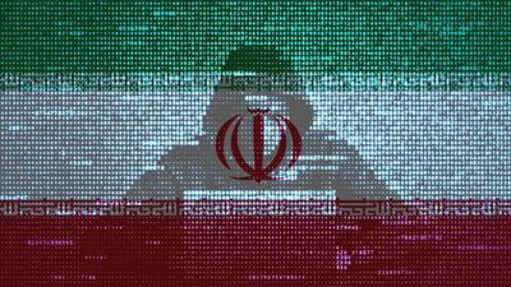 U.S. Needs To Upgrade Critical Infrastructure To Counter Iranian Hackers A conceptual digital illustration features a pixelated Iranian flag overlaying the silhouette of a hooded figure operating a computer terminal, symbolizing cyber threats posed by Iranian hackers.