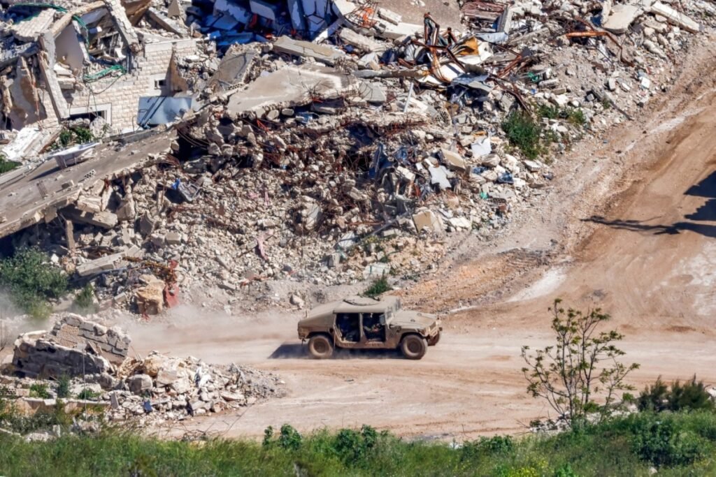 A military Humvee maneuvers along a dusty, unpaved road adjacent to heavily collapsed concrete structures and piles of building rubble in Lebanon, providing a visual illustration of the regional devastation linked to an incoherent strategy.