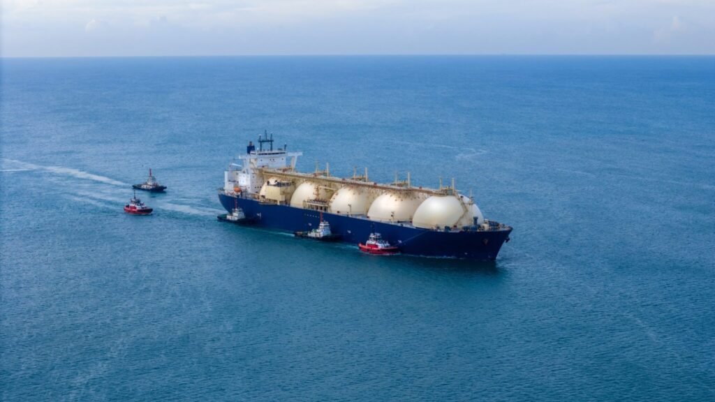 The LNG Security Myth: How the Strait of Hormuz Exposes LNG’s Vulnerabilities A large LNG carrier with spherical storage tanks being escorted by several small red tugboats across a calm blue sea, illustrating the logistical complexity and infrastructure dependency of the Global LNG supply amid severe geopolitical challenges.