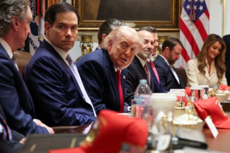 Donald Trump leans forward at a crowded cabinet table alongside Marco Rubio and other political advisers with red hats scattered in the foreground, visually highlighting the strategic challenges surrounding Trump's missions on global foreign policy.