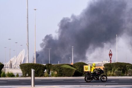 A delivery driver riding a motorcycle past manicured roadside shrubbery while thick black smoke rises into the desert sky from a distant facility explosion, contextualizing the physical disruptions to tech infrastructure under The Impact of the Iran War.