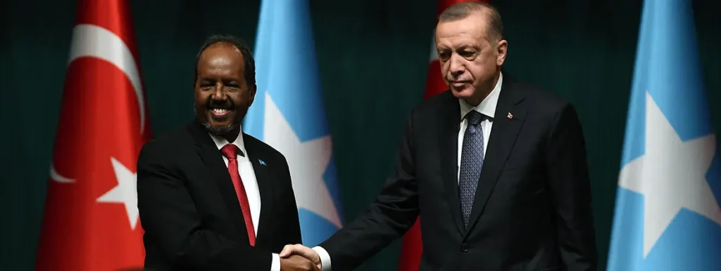 Turkish President Recep Tayyip Erdoğan and Somali President Hassan Sheikh Mohamud shaking hands in front of their national flags, representing the bilateral cooperation behind the Turkish Somali hydrocarbon geostrategy.