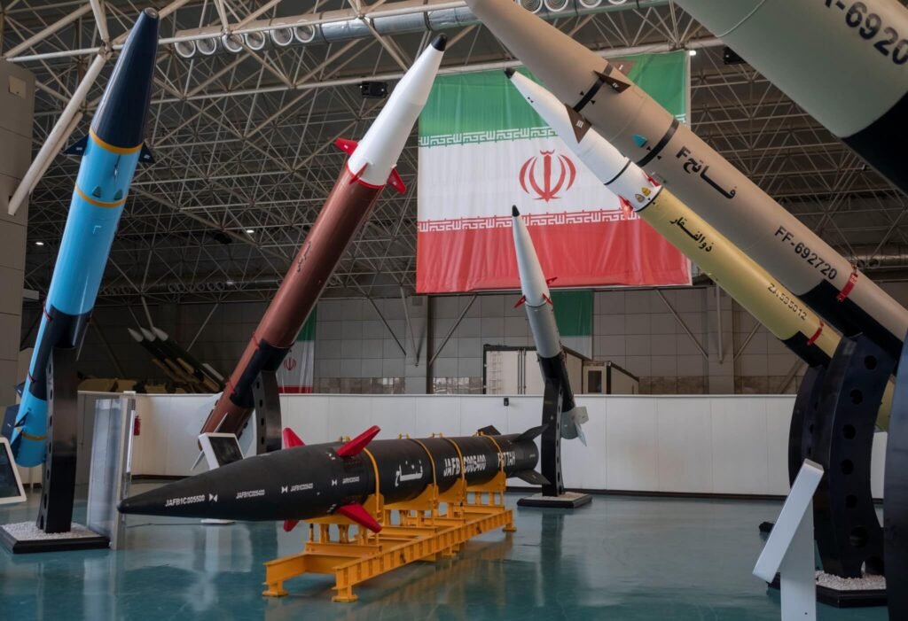 ارزیابی خسارات به زرادخانه موشکی بالستیک ایران An indoor military exhibition showcasing various precision-guided weapons and defense systems on launch cradles under a large Iranian flag, serving as a primary visual for evaluating the damage to Iran's ballistic missile arsenal.