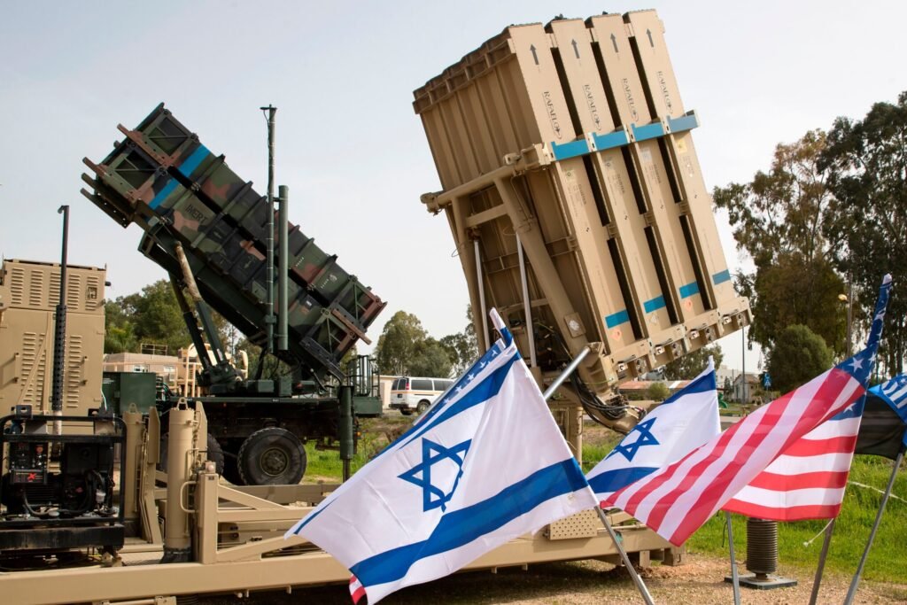 An outdoor deployment of an Iron Dome missile defense battery alongside a Patriot missile system, flanked by prominent Israeli and US flags, visually representing the strategic defense hardware linked to discussions on a US military aid drawdown.