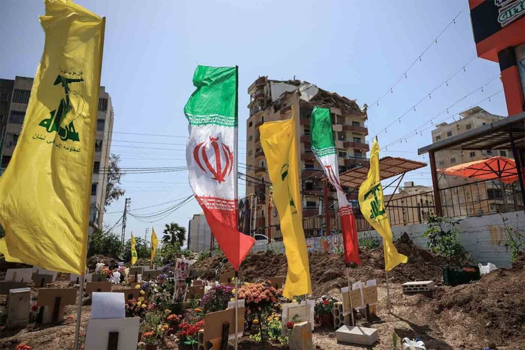 Yellow Hezbollah flags and Iranian national flags fly prominently over an outdoor memorial and burial site with floral tributes, set against damaged residential buildings to illustrate the regional reach of the Iranian Proxy Network.