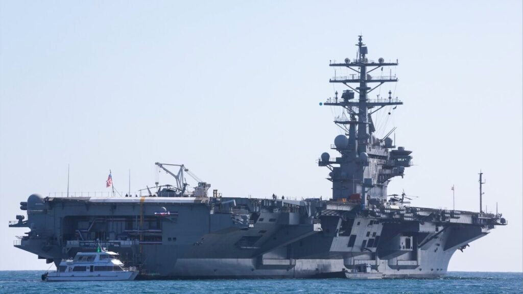 A massive aircraft carrier at sea with a smaller support vessel alongside, representing the naval force and international pressure involved in the Iran blockade regime collapse strategy.